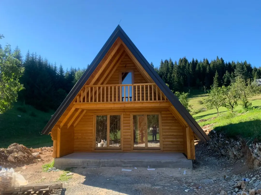 Wooden A-frame house with a balcony on the gable, below a forest