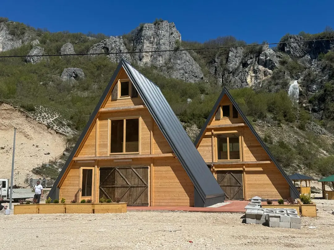 Dvije A-frame kuće od drveta sa tamnim limenim krovom ispod stijena